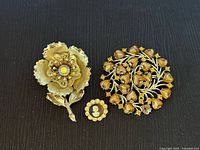 Front view of three vintage brooches on black fabric background showing detailed floral, heart leaf, and cameo designs.
