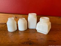 Four white milk glass jars displayed side by side on a wood surface against a red wall, showing their varying shapes and sizes without lids.