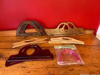 Full view of 6 assorted purse handles displayed on wooden surface against red background, showing variety of wood finishes, acrylic, and sealed plastic set.