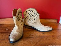 Pair of tan suede cowboy boots with mixed metal studs and faux gems on a wood floor against red background. Side and angled views show the condition and embellishments clearly.