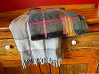 Two throw blankets folded on a wooden cabinet showing contrasting neutral plaid and colorful plaid with fringe edges.