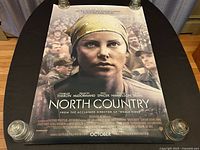 Front view of the double-sided North Country movie poster laid flat on a table, held at corners with glass weights. Shows close-up image of Charlize Theron with a yellow bandana and crowd in the background.