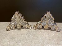 Pair of matching silver-plated napkin holders with a shell and scroll motif on a stone countertop background under warm lighting.