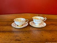 Pair of vintage tea cups and saucers on wooden surface against red background. Left cup is white with gold starburst and red detail (Paragon); right cup is cream with gold wheat motif (Shelley Golden Harvest).