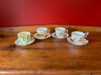 Four vintage English bone china teacups and saucers displayed on wooden surface with red background, showing varied floral designs and shapes.