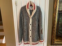 Full view of the Roots Cabin cardigan hanging, showing marled grey knit with cream trim, button front, red stripe accents, and elbow patches.