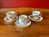 Three vintage bone china teacup and saucer sets together on wooden table showing distinct floral patterns and different shapes.