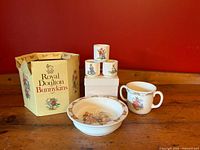 Photo showing the two-piece Bunnykins baby set with double-handled cup and baby plate, three extra Bunnykins cups stacked, and the original hexagonal box showing wear.