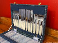 Front view of open navy blue polka dot case holding six fish forks and six spreader knives vertically in upper tier, showing faux mother-of-pearl handles and shiny chrome-plated blades and tines.