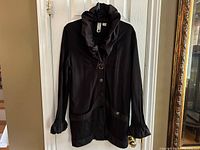 Full front view of black longline jacket hanging on door, showing ruffled trim on collar and cuffs, and snap buttons down front with oversized pockets.