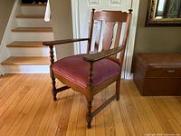 Side-angle view showing the turned wooden legs and arms and the pink velour upholstery on the seat, situated on a hardwood floor with stairs visible in the background.