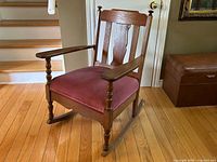 Side angled view of the oak rocking chair showing turned spindle legs and armrests, pink velour seat, and decorative backrest.