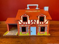 Front view of the Fisher-Price Tudor-style dollhouse showing red roof, blue handle on top, blue front door, and detailed lithographed brick and timber design with window flower boxes.