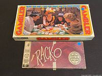 Boxes of Gambler and Rack-O vintage board games side by side on black surface, Gambler box top shows people playing casino style game, Rack-O box purple with gold seal