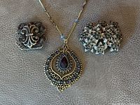 Photo showing three vintage gothic-inspired jewelry pieces: a large drop-style filigree necklace with a red faceted teardrop stone, and two brooches featuring black rhinestone flowers and scrollwork design.