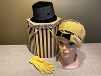 Front view of the yellow veiled straw hat displayed on a mannequin head, black velvet pillbox hat, yellow gloves, and striped vintage hat box.