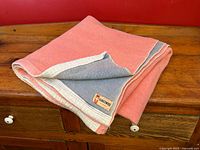 Vintage Kingswool wool blanket folded on wooden surface showing pink side and faded blue reverse with cream satin binding.
