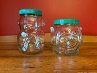 Front view of two clear glass Kraft peanut butter jars shaped like bears with green lids, on a wooden surface with a red background.