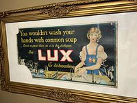 Gold-tone ornate framed vintage Lux soap advertisement featuring a woman washing dishes with text promoting Lux for dishwashing, visible wear and tears on paper ad.