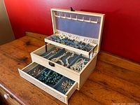 Open vintage Mele jewelry box showing top tray with sections containing rings, brooches, and cufflinks, middle compartment with beaded necklaces, and lower drawer with additional jewelry.