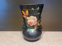 Front view of the matte black ceramic vase showing the hand-painted pink rose, orange and yellow buds and green leaves on the ridged sculptural body with bulbous base