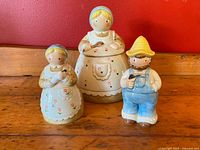 Front view of all three ceramic pieces on wooden surface with red background, showing detailed country farmhouse figurines.