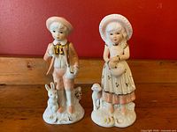 Front view of both porcelain figurines, a boy with a rabbit and a girl with a lamb and dog, on a wooden surface against a red background.
