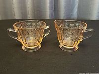 Both pink Depression glass pieces side by side on a black surface, showcasing their matching floral scroll patterns and handles.