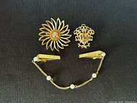 Top-down view of three pieces: floral brooch, ornate baroque-style brooch with amber glass, and sweater guard clip with faux pearl chain.