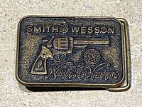 Top view close-up of the vintage Smith & Wesson brass belt buckle with detailed raised revolver and embossed logo, showing surface texture and patina.