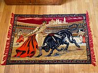 Full view of the vintage Spanish bullfighting tapestry on wood floor showing the entire tapestry with red fringe and detailed bullfight scene.