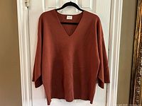 Front view of the oversized rust colored Wilfred Free sweater hanging on a black hanger, showing the full body of the sweater, V-neckline, and drop shoulder sleeves.