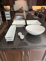 Full set of assorted white ceramic serving dishes arranged on a kitchen island including bowls, rectangular plates, square bowls, and small cups.