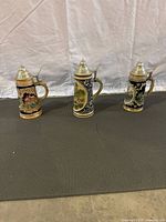 Front view of all three beer mugs lined up showing their different illustrated designs and pewter lids.