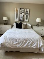 View of two white bureaus or bedside tables positioned on each side of a bed, each with three drawers and round knobs, placed against a beige wall with decorative feather artwork above. One drawer has repaired damage.