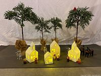 Full set of five lit ceramic village houses with four tall artificial Christmas trees, two small clear animal figurines, two Russian nesting dolls, and two black horse figurines decorated with wreaths, arranged on gray fabric.