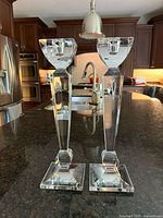 Front view of both candlesticks showing their height, clear lead crystal material, and square bases on a dark granite countertop with kitchen background.