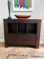 Front view of the dark stained oak sideboard with three open cubbies on top and three cupboard doors below, displaying two decorative items on top.