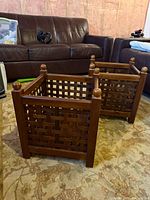 Two wooden square planter boxes with woven lattice sides and round finials on the four corner posts, displayed on a patterned carpet with a brown leather sofa in the background.