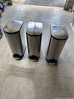Three identical tall stainless steel step garbage bins with black plastic bases, liners, and split butterfly lids, seen from the front.