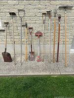 Row of ten gardening tools lined up against brick wall showing full length of handles and heads