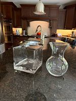 Photo showing both glass flower vases on a kitchen counter with dark granite surface, wood cabinets and kitchen appliances in background.
