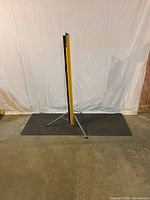 Vintage portable projection screen rolled inside a yellow cylindrical case, standing upright on tripod legs on a floor with white backdrop.