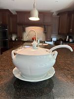 White porcelain soup tureen set shown on kitchen countertop, depicting tureen with lid and ladle inside the tureen and a plate underneath.