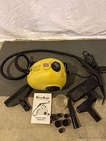 Yellow Steam Buggy steam cleaner with steam hose, several attachments, five small brushes, measuring cup, and manual laid out under white sheet background.