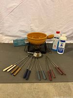 Full kit including stoneware crockery pot on metal stand with burners, 12 fondue forks, fuel bottles, and heat diffusers on gray mat