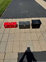 Three tool boxes closed, red steel (left), grey steel (middle), black plastic (right), arranged on paved surface.