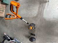 Orange and gray Worx battery powered whipper snipper with attached Power Tank battery, shown fully assembled on concrete floor.
