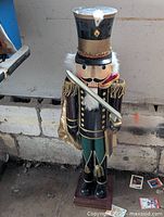Full view of the 34 inch wooden nutcracker soldier showing black and gold uniform, green pants, white hair and beard, and sword held across the chest. Base visible. Condition shows need to glue legs.
