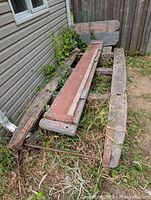 Antique 18th century wooden Shugart shack sled, showing overall view with heavy oak beams, iron reinforcements, and reddish-faded central plank, resting on ground near a building.